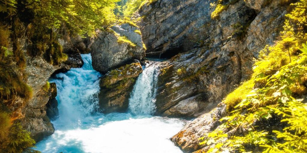 Klares, türkisblaues Wasser fließt durch eine felsige Schlucht inmitten einer grünen, sonnendurchfluteten Landschaft.