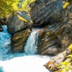 Klares, türkisblaues Wasser fließt durch eine felsige Schlucht inmitten einer grünen, sonnendurchfluteten Landschaft.